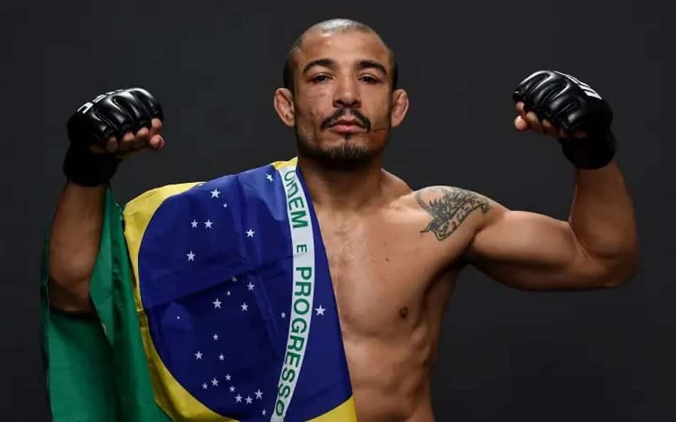 Com a bandeira do Brasil sobre o ombro direito, José Aldo, de luvas, mostra os bíceps e faz expressão série em sessão de fotos do UFC