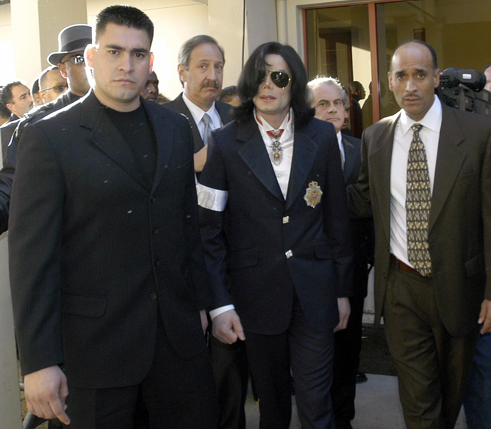 Michael Jackson exits a courthouse in dark sunglasses, a black suit, and a medal around his neck, flanked by his lawyer Mark Garagos and an entourage.