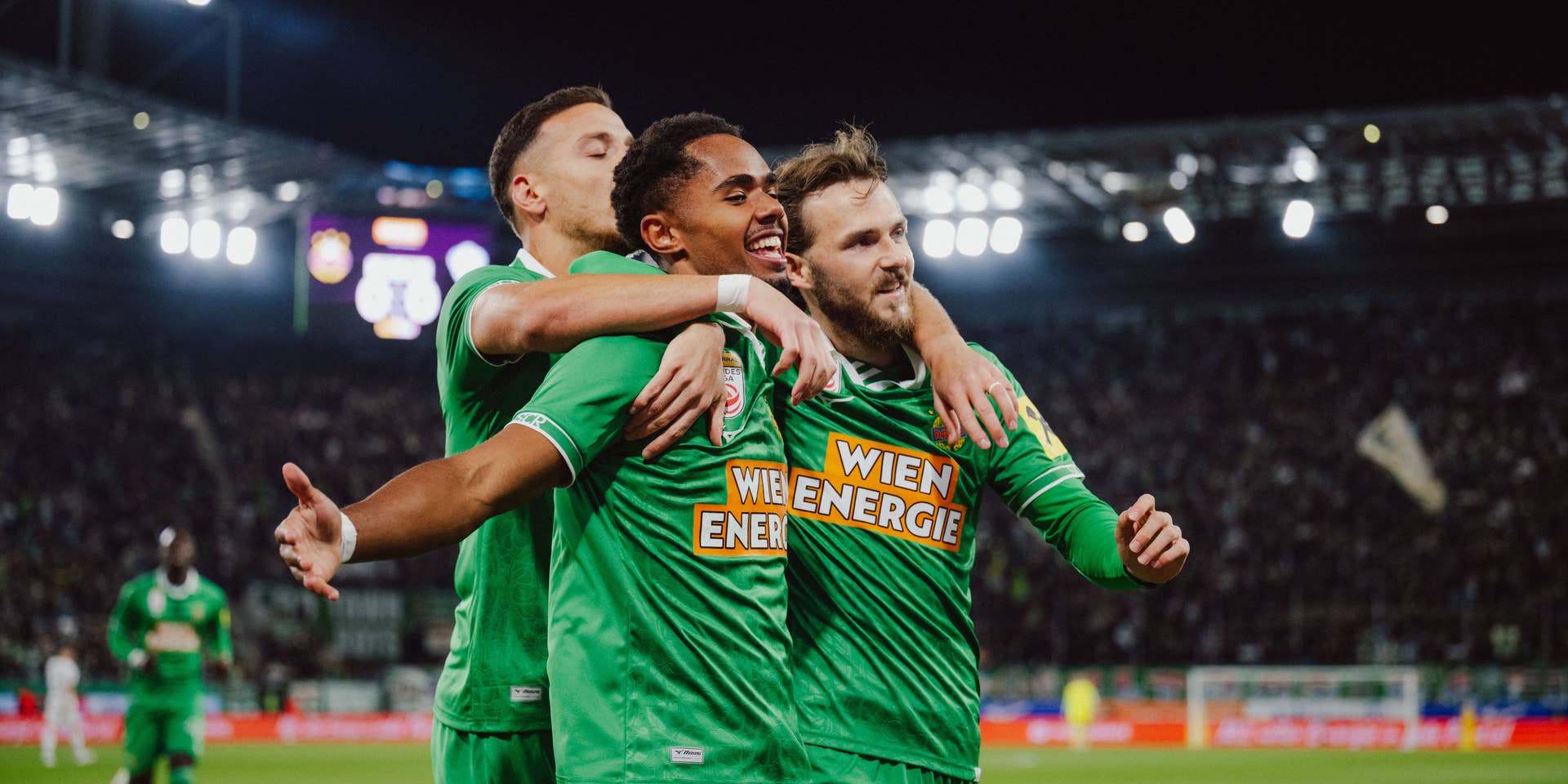 Petter Dahl jubelt strahlend im grünen Trikot und wird von seinen Rapid-Teamkollegen herzhaft umarmt.