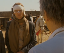 Owen Wilson with a bandage over his eyes, talking to another person with horses and a rustic background
