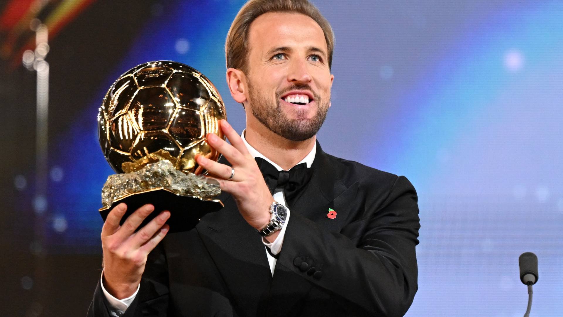 Homem branco, barba rala, cabelo loiro, sorrindo e segurando um troféu dourado em formato de bola de futebol. Ele veste um terno preto com gravata borboleta e um broche de papoula vermelha na lapela, em um fundo azul iluminado