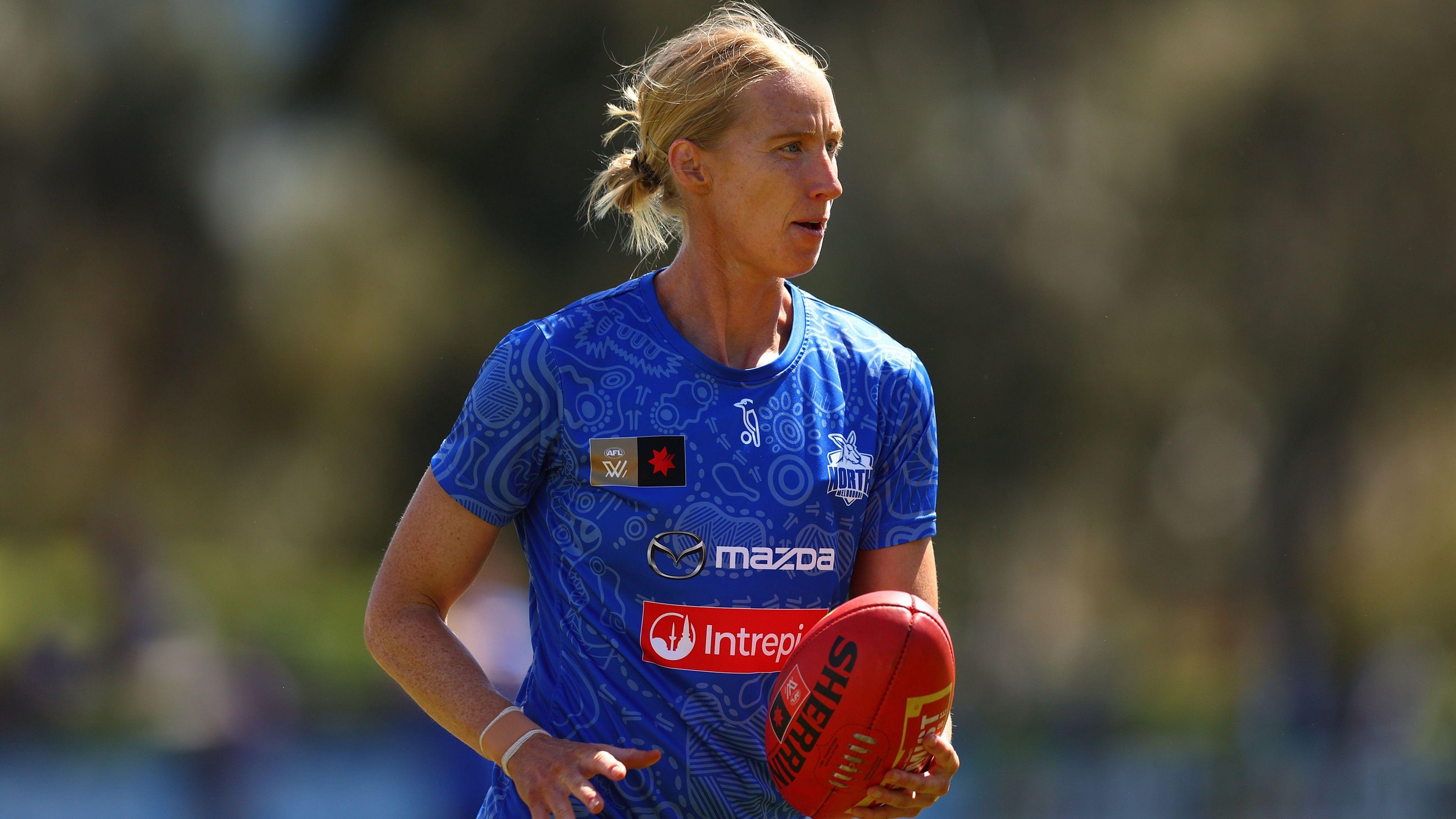 A player in a blue Australian rules football jersey holds a red football while on a sports field. The player is pictured from the waist up, with a blurred outdoor background and sponsor logos visible on the shirt.