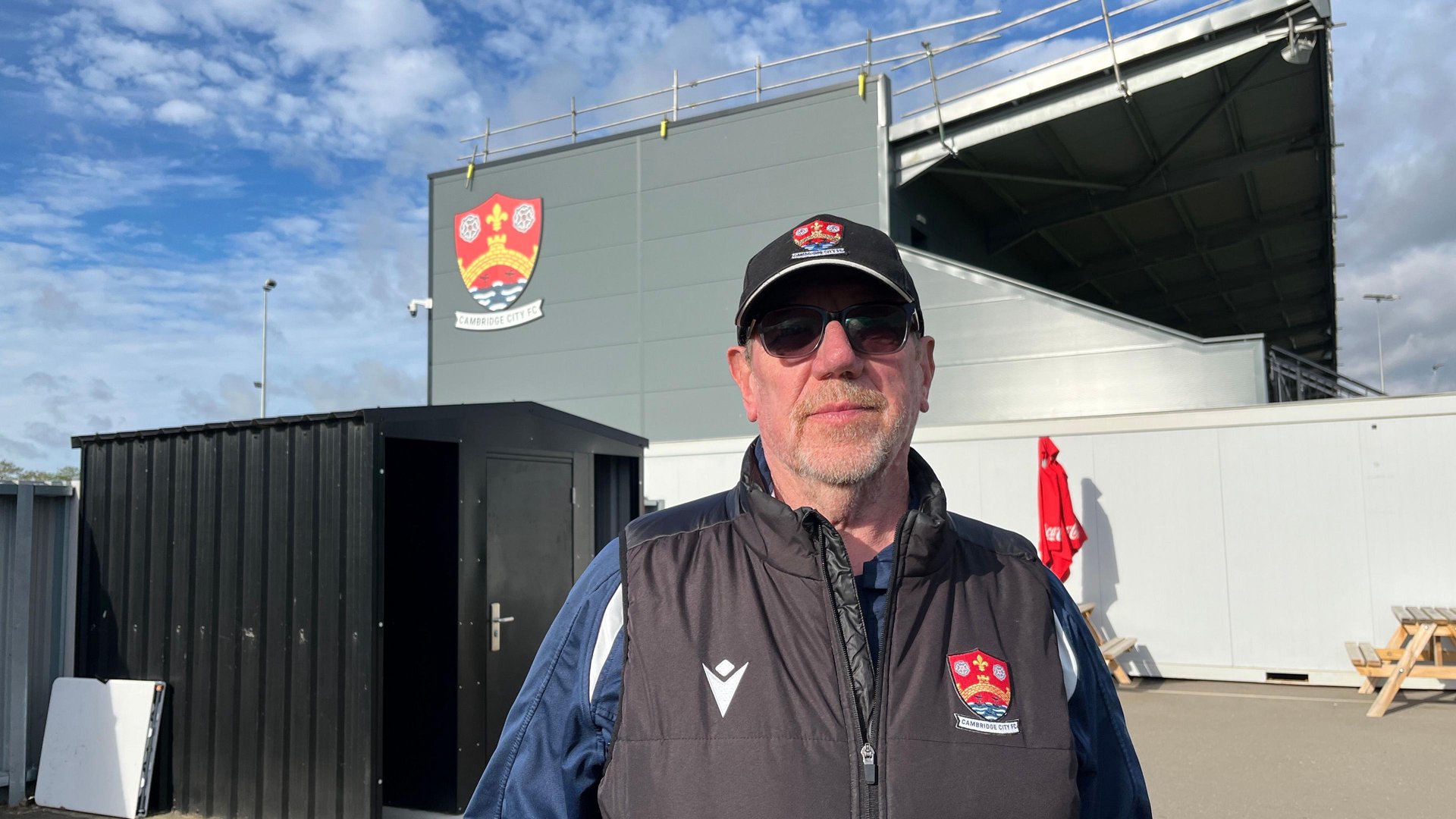 Phil Brasher is standing in front of a football stadium. He is wearing a blue football shirt and a branded gilet as well as a baseball cap.