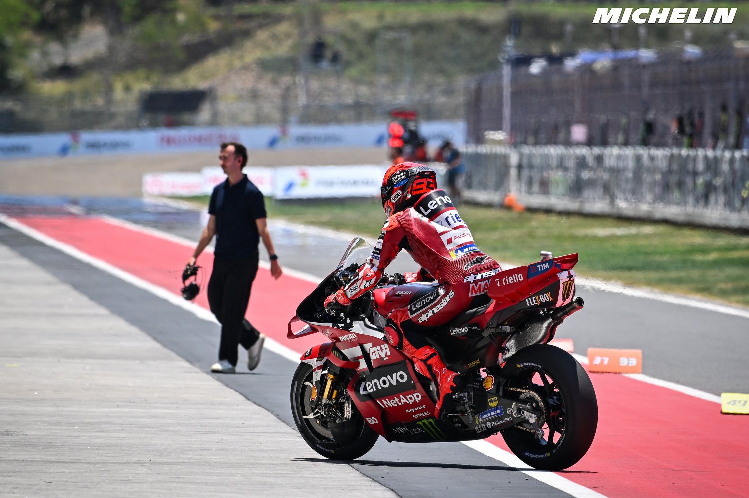 Marc Marquez on his red Ducati Desmosedici with number 93 leaving the MotoGP track, watched by a man in black walking nearby