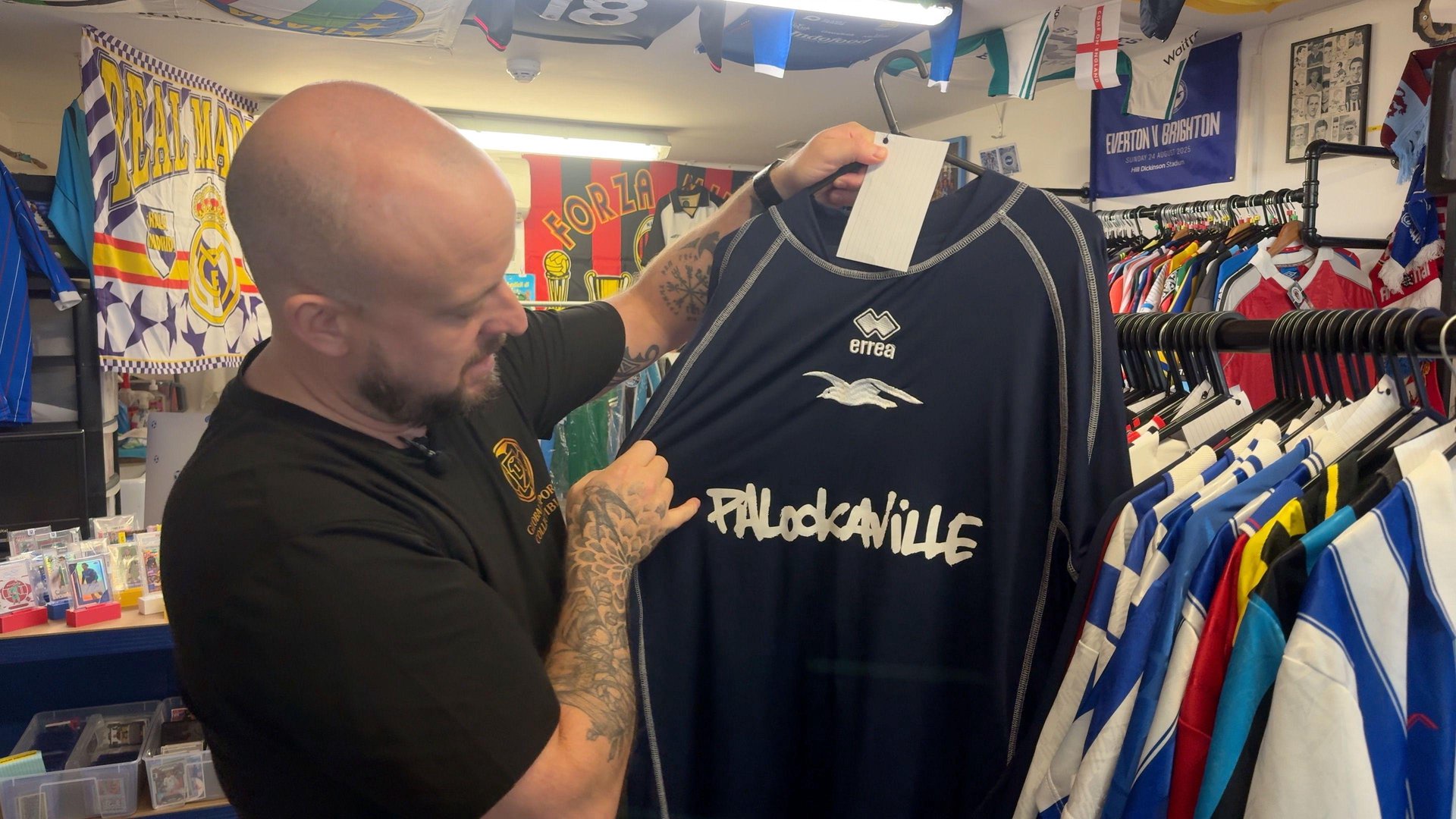 A bald man with a black t-shirt and tattoos on his arm is holding up a black football shirt to the camera. It is 'Palookaville' written on it in white, with a white seagull printed above. To the right of the shirt, are dozens of other football shirts hanging up