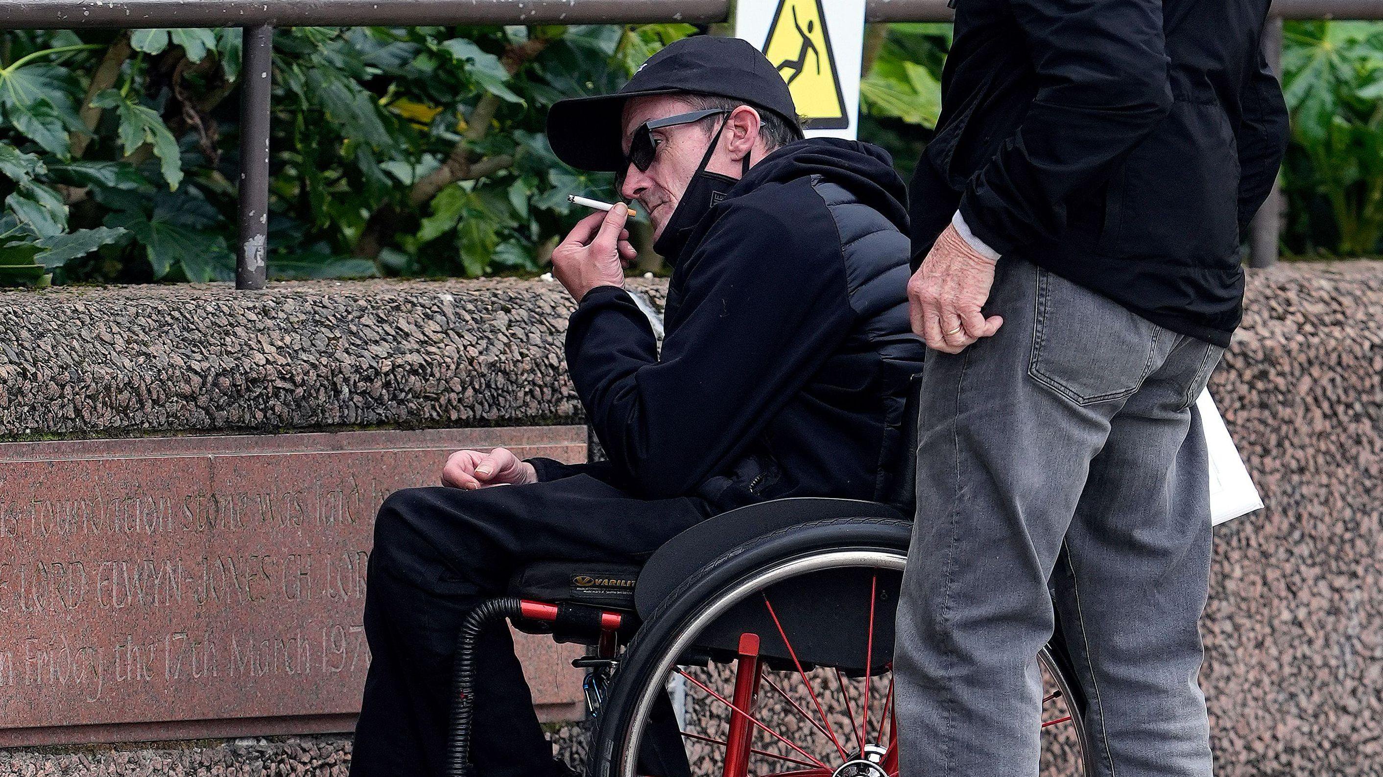 A man in a wheelchair wearing a dark navy hooded jacket with a quilted front is photographed holding a ciggarette towards his mouth. He is wearing dark sunglasses.