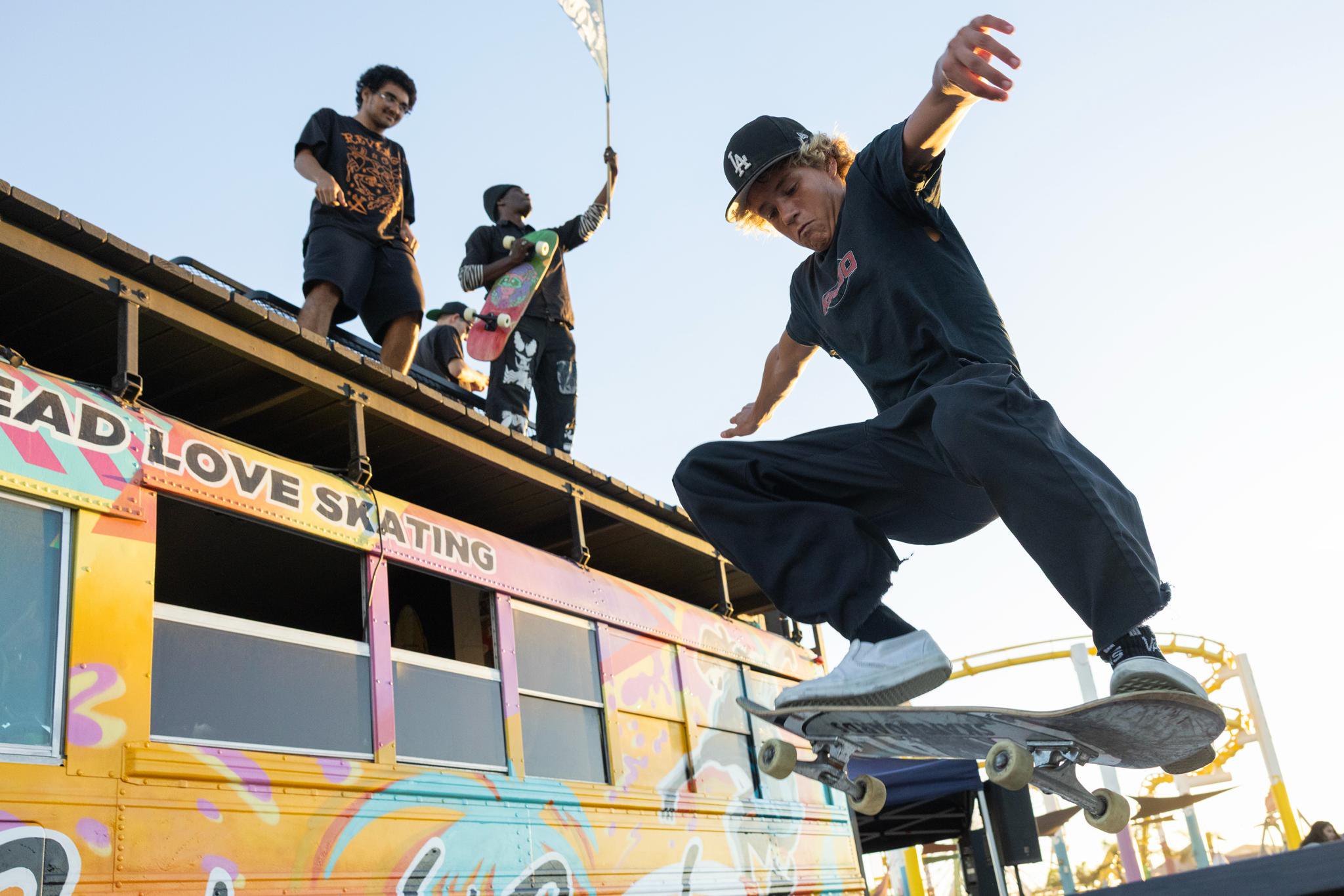 A skateboarder performs a trick in the air near a colorful bus decorated with the words "Spread Love Skating" during Locals Night at the Santa Monica Pier.