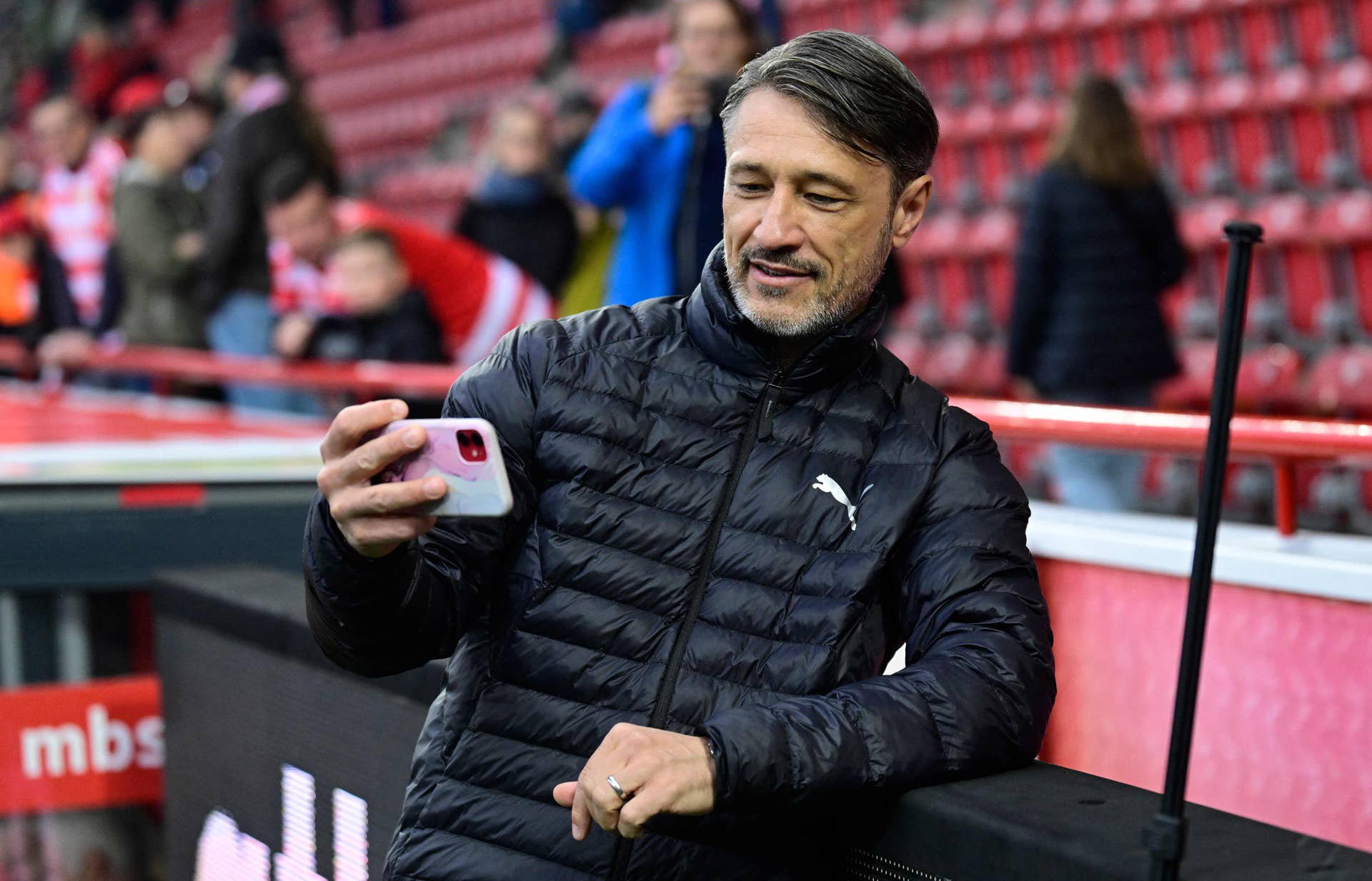 Niko Kovac steht in einer schwarzen Daunenjacke im Stadion an der Bandenwerbung und blickt schmunzelnd auf sein in der Hand gehaltenes Smartphone.