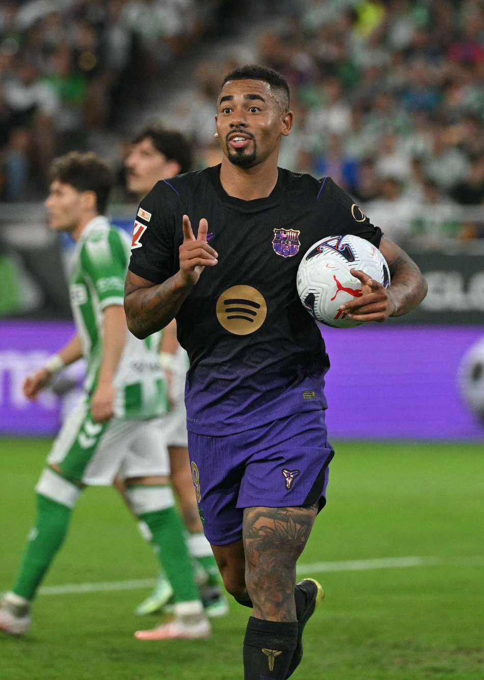 Gabriel Jesus, jogador do Barça, corre com a bola durante partida, com torcedores ao fundo.