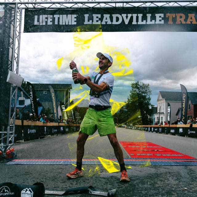Runner celebrating with champagne after finishing the Leadville Trail race, surrounded by spectators and banners.