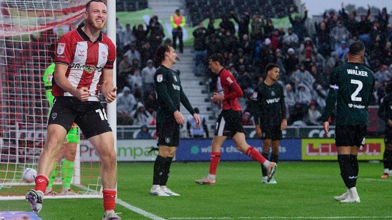 Lincoln City player Ben House, in red and white stripes, grins and celebrates his goal against Burnley, with the ball in the net in the background and three Burnley players in all black standing disconsolately.