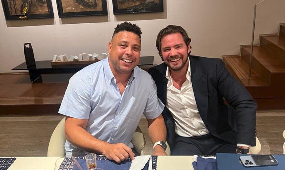 Ronaldo Fenômeno, de camisa azul clara, e um homem de terno escuro e camisa branca sorriem para a câmera, sentados à mesa. Ao fundo, quadros e uma escada de madeira com corrimão de vidro