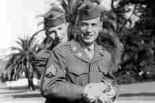 Two male soldiers wearing WWII-era formal uniforms embrace in this black-and-white photograph. Palm trees are visible in the background.