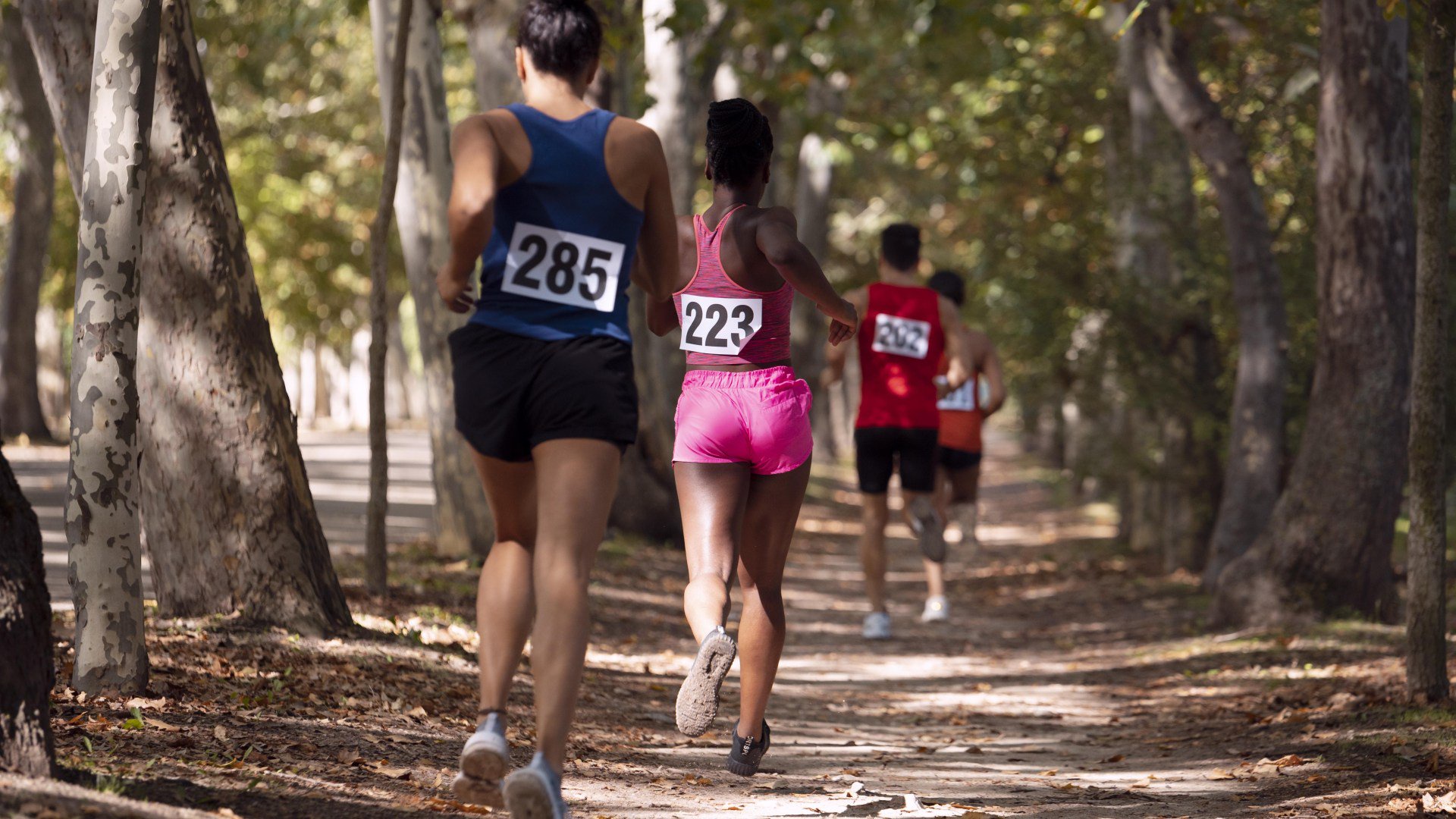 Corredores em uma trilha de terra arborizada. Uma mulher de regata azul com o número 285 e shorts pretos lidera, seguida por outra de regata rosa com o número 223 e shorts pink. Outros corredores estão mais ao fundo.