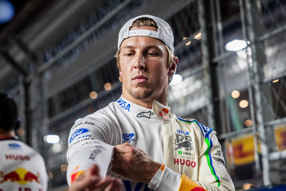 A Red Bull Racing driver in a white race suit with Visa Cash App and Ford logos stands in the paddock, adjusting his glove near a pit wall.