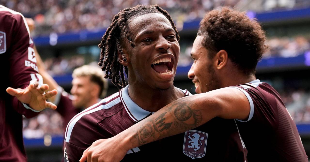 Switzerland footballer Johan Manzambi smiling and being embraced by teammates to celebrate a goal while playing for Premier League team Aston Villa Football Club in a claret shirt with light blue trim and the club badge and Adidas logo on his chest