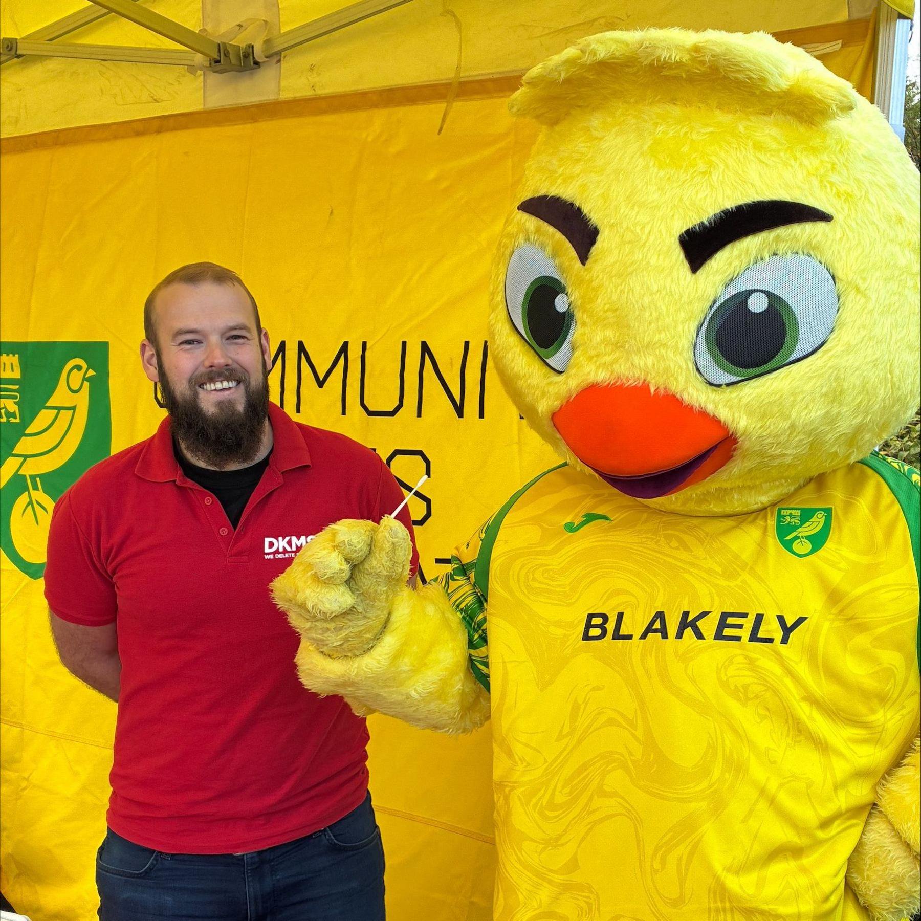 A man who works for a blood cancer charity is wearing a red T-shirt and is standing on the left of the image. On the right is a mascot of a canary with an orange beak, big green eyes and dark eyebrows. It is wearing a yellow and green Norwich City Football Club T-shirt. The mascot is holding a long cotton swab in its hand. 