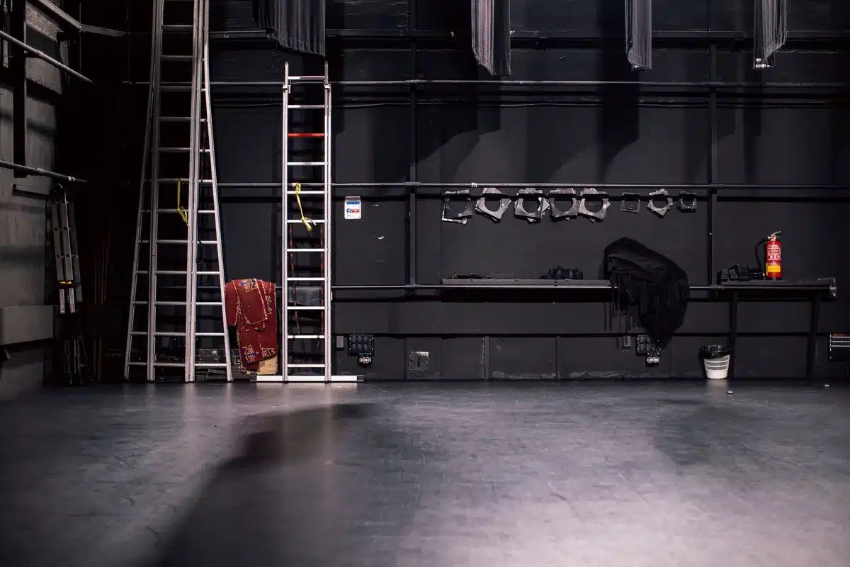 Empty West End theatre stage with a single spotlight on a golden orb on a small pedestal, red velvet curtains framing the scene