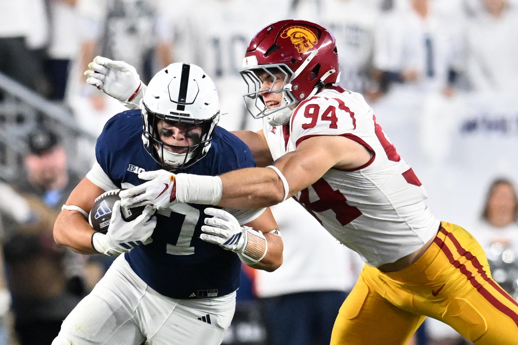 A Penn State football player in a blue uniform carries the ball while being tackled by a USC player in a white and maroon uniform with the number 94.