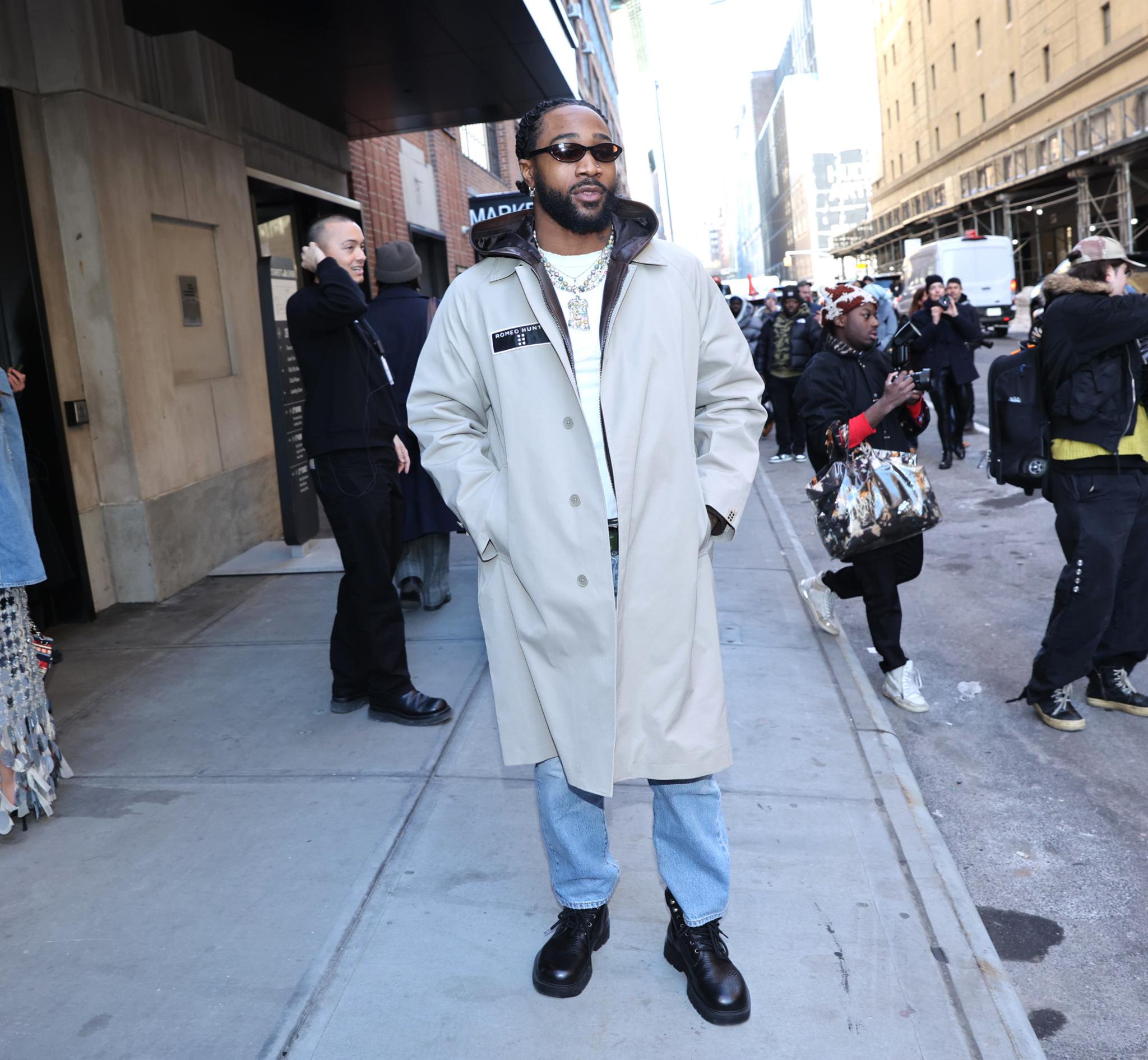Miles Sanders arrives at a New York Fashion Week event, wearing a tan trench coat, layered hoodie, and sunglasses, on a New York City sidewalk.