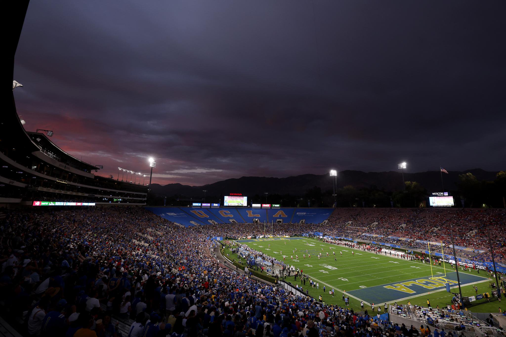 Wide-angle view of a packed football stadium under a dramatic, dark sky at dusk.