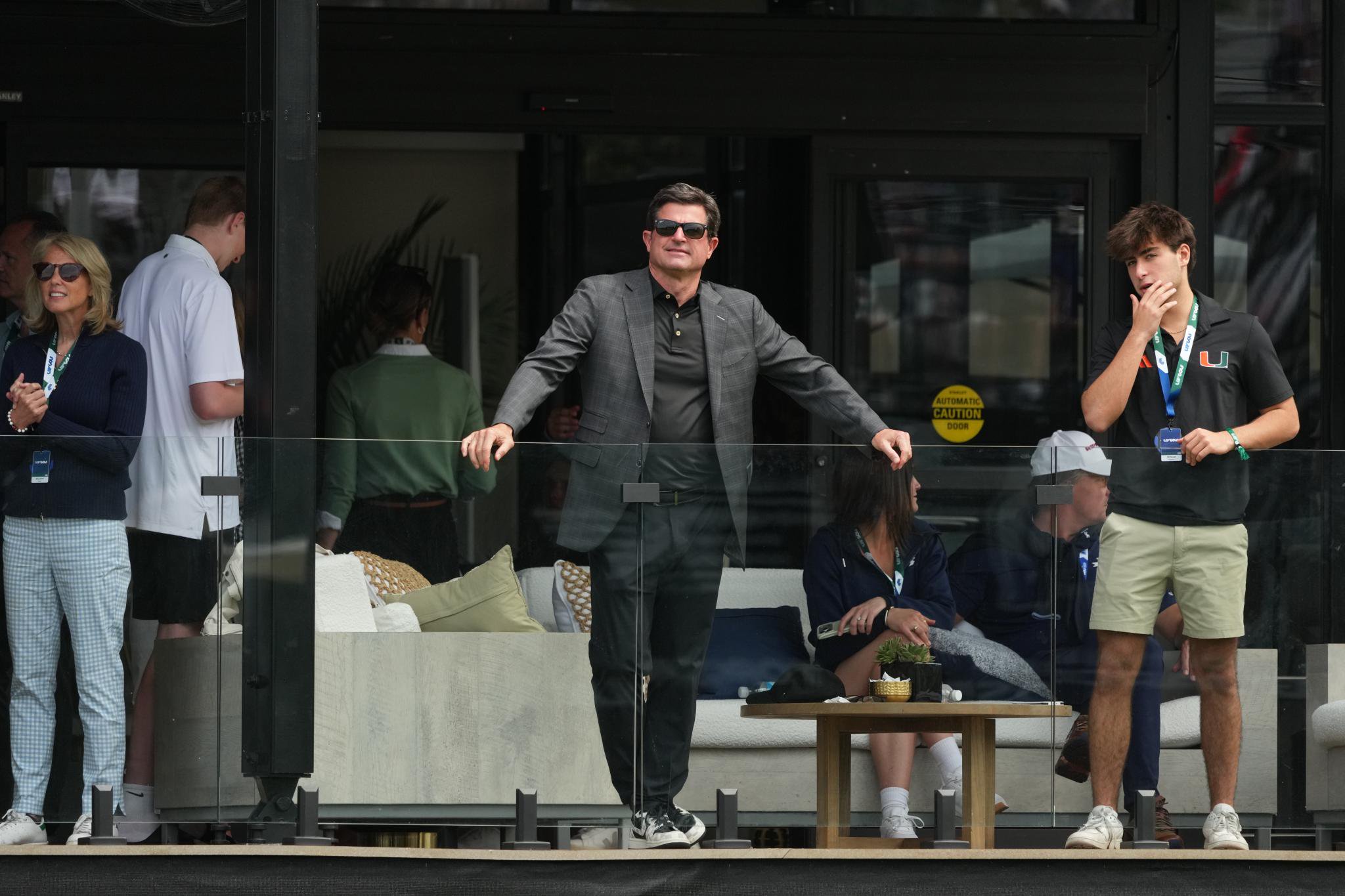 LIV Golf CEO Scott O'Neil stands on a balcony overlooking a tournament, flanked by spectators.