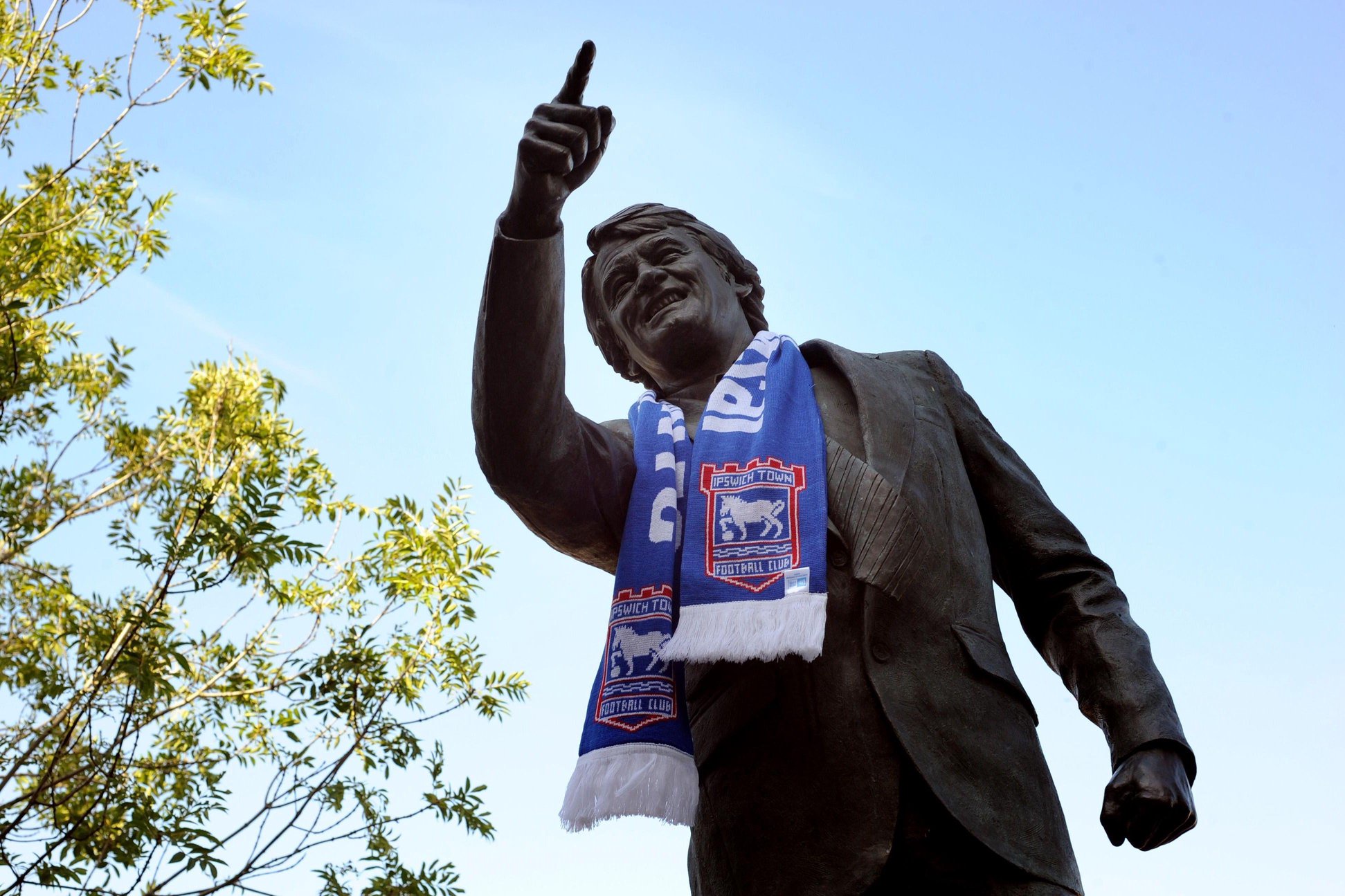 A bronze statue of Sir Bobby Robson, who is depicted pointing and smiling. The statue is pictured from the ground looking up at it. There is a blue Ipswich Town scarf draped round his neck. The sky is blue above.