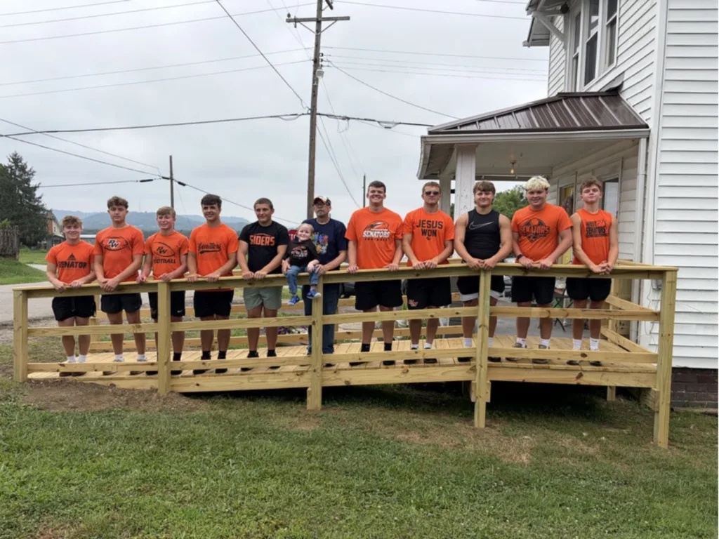 Players in orange football shirts lean on the railing of a new wooden ramp with Maverick outside a white house