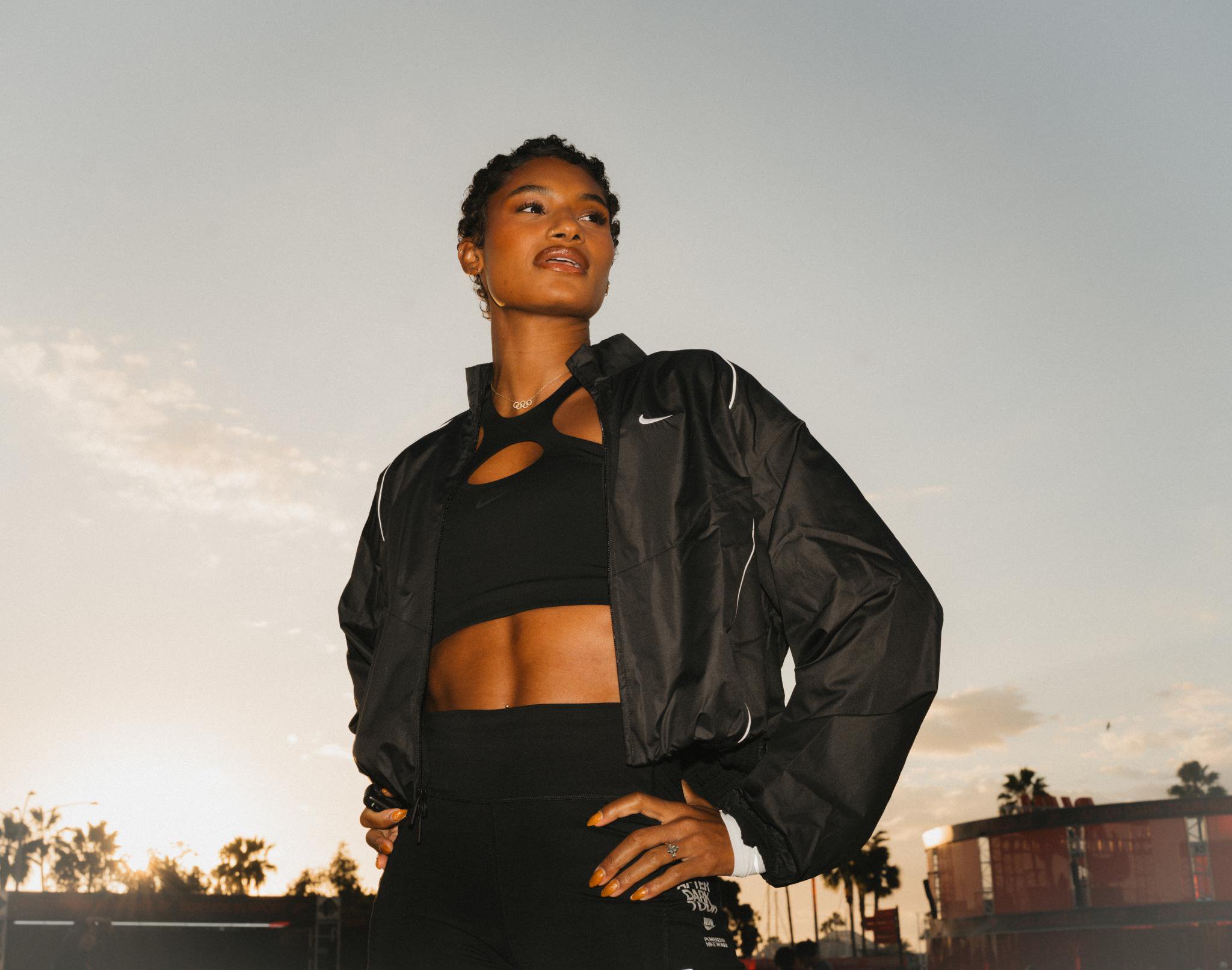 A Black woman in athletic wear looking ahead, with hands on her hips, against a dusky sky.