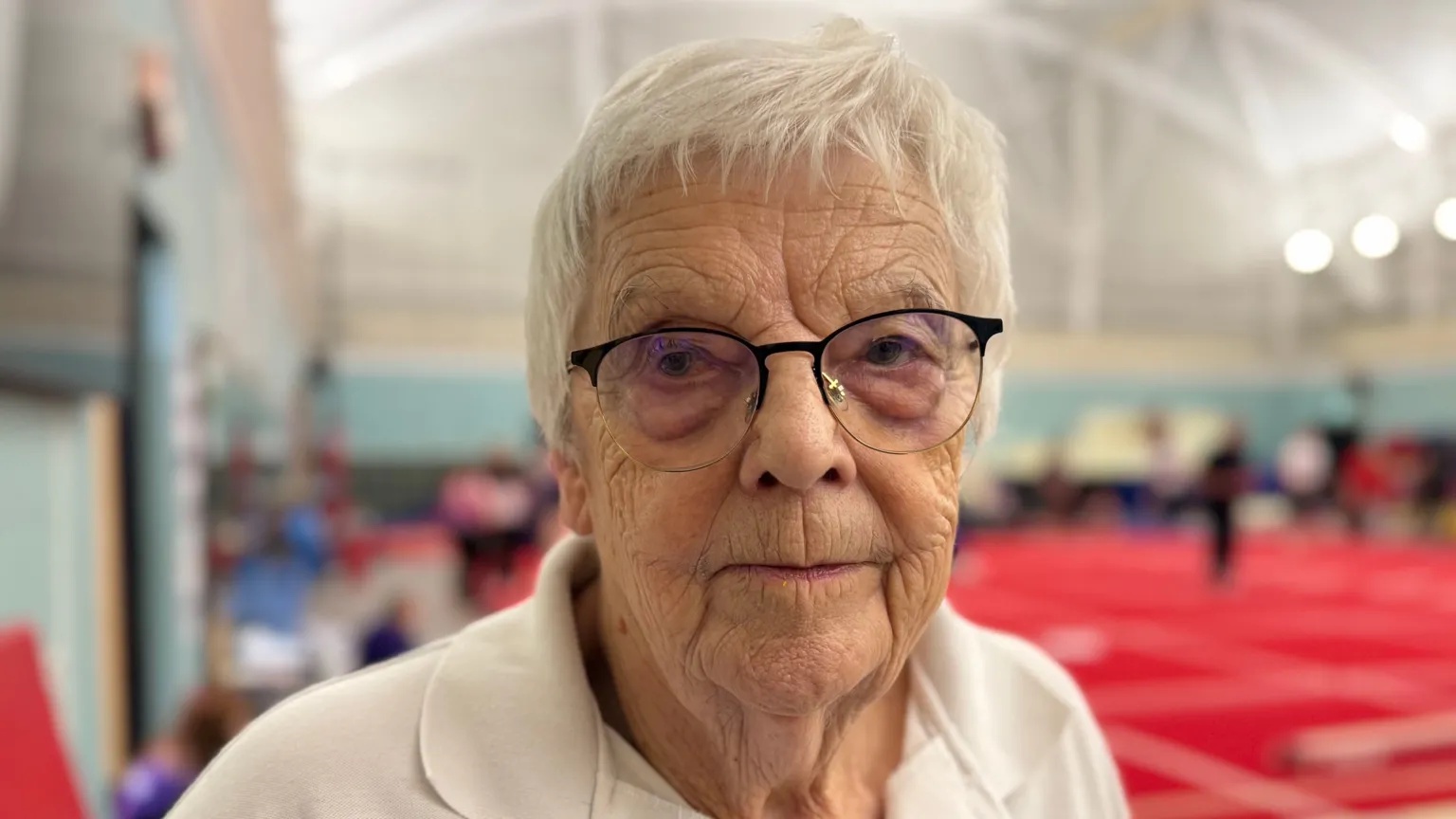 A close-up of Kay Salter in a gym. She has a white polo shirt and has short white hair and glasses.