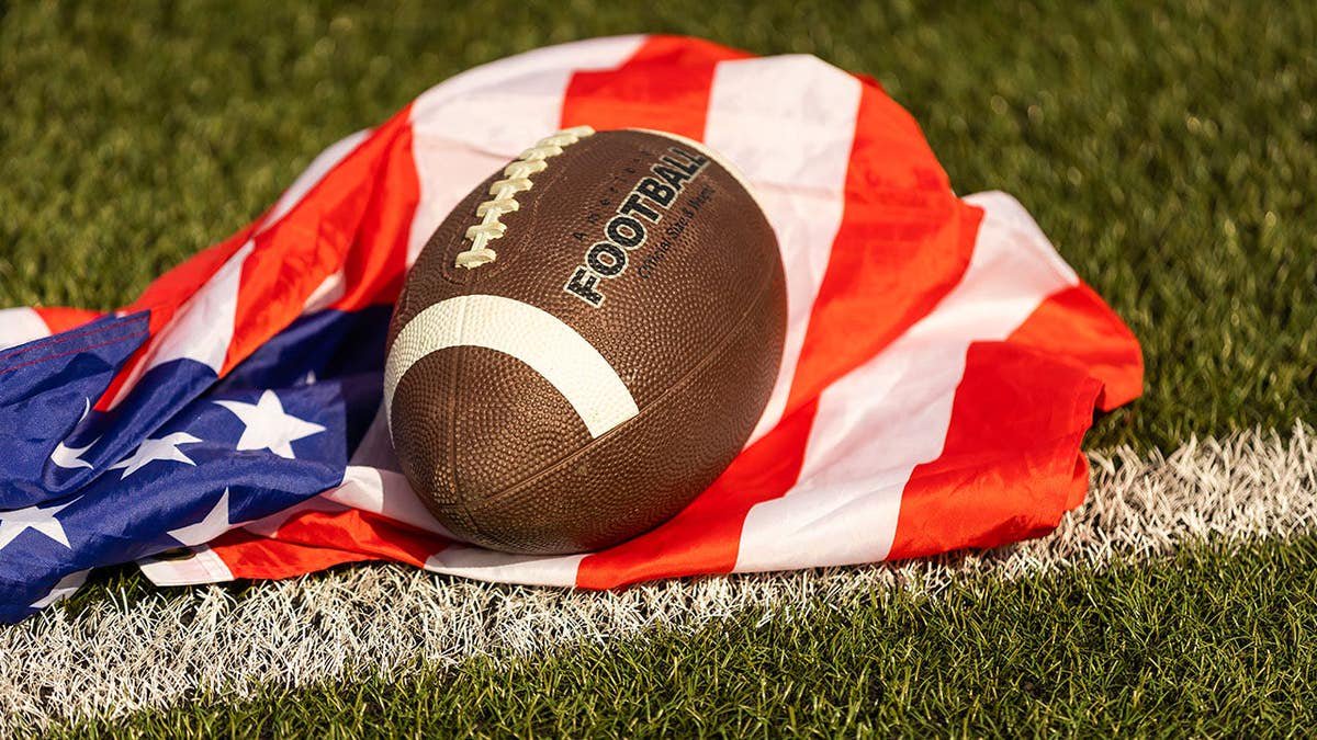 American football rests on green grass with a U.S. flag background.