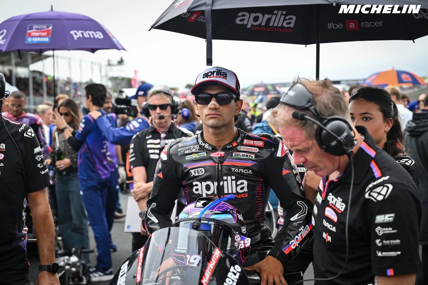 A rider in black Aprilia Racing leathers and cap on the MotoGP grid, with crew in headsets beside him and umbrellas overhead.