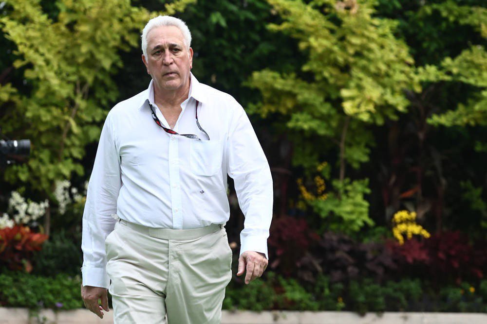A white-haired man in a white shirt and beige trousers walks outdoors past green foliage and flowering plants, with sunglasses hanging from his shirt pocket.