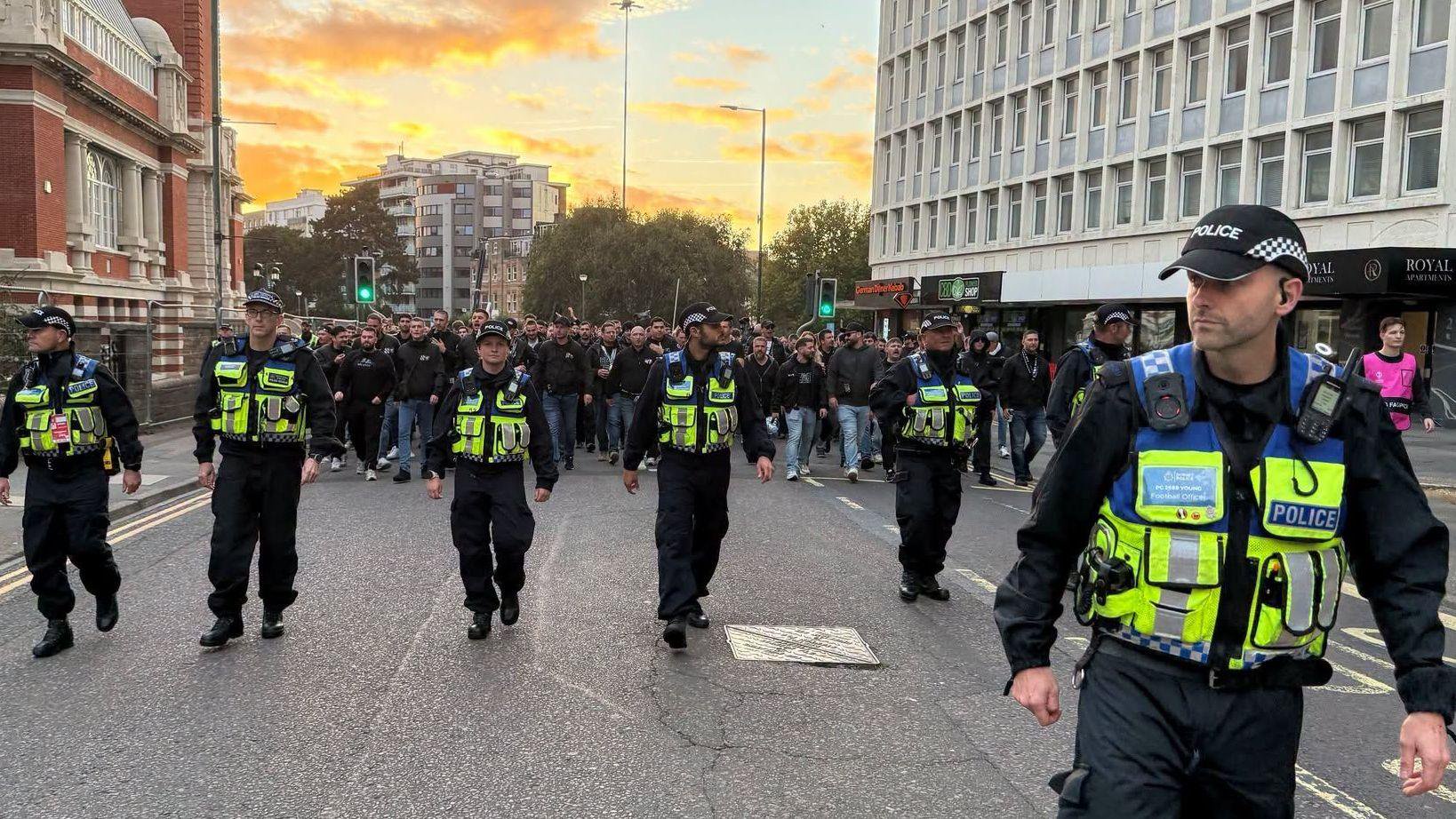 Police officers in a line with large crowd of visiting football fans in Bournemouth - it is early evening with a golden sunset. 