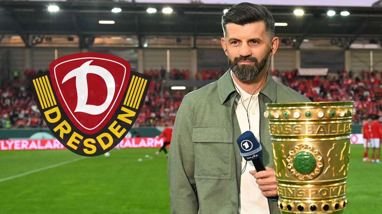 Schalke-Trainer Miron Muslic mit einem ARD-Mikrofon in der Hand steht hinter der DFB-Pokal-Trophäe und schaut auf das Vereinslogo von Dynamo Dresden.