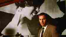 Kevin Costner in The Bodyguard with a picture of Whitney Houston behind