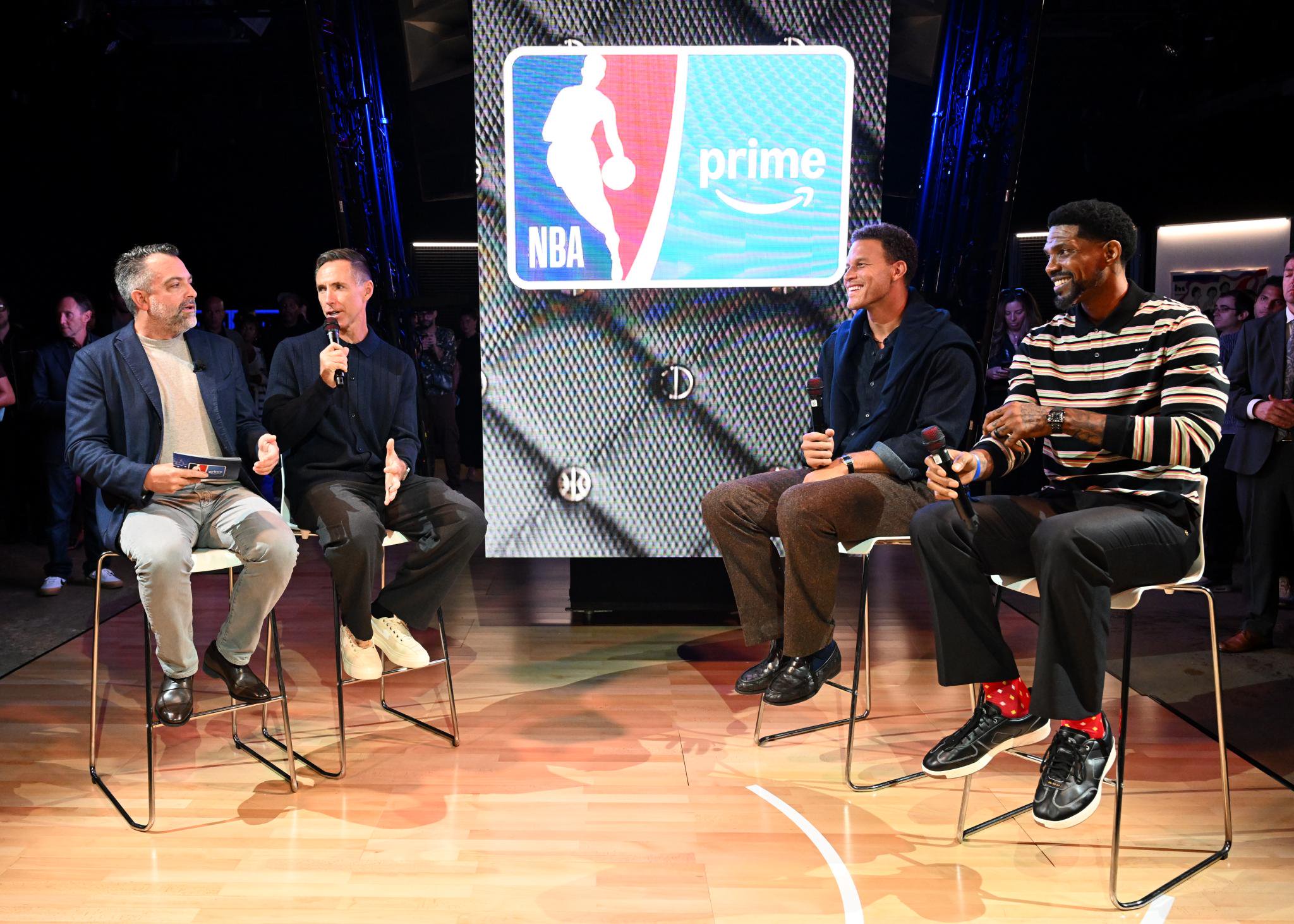 Four men sit on high stools on a stage during the NBA on Prime season 2 launch event, with a large screen behind them displaying the NBA and Prime logos.