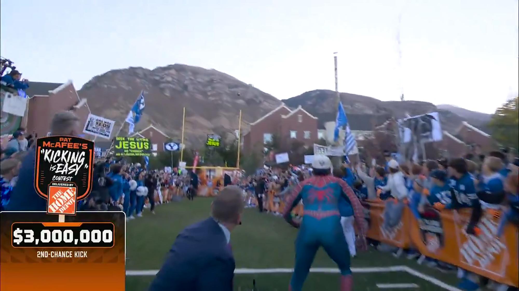 A kicker dressed in a Spider-Man suit prepares to kick a field goal during the Pat McAfee Show kicking contest, watched by a large crowd in front of a mountain.