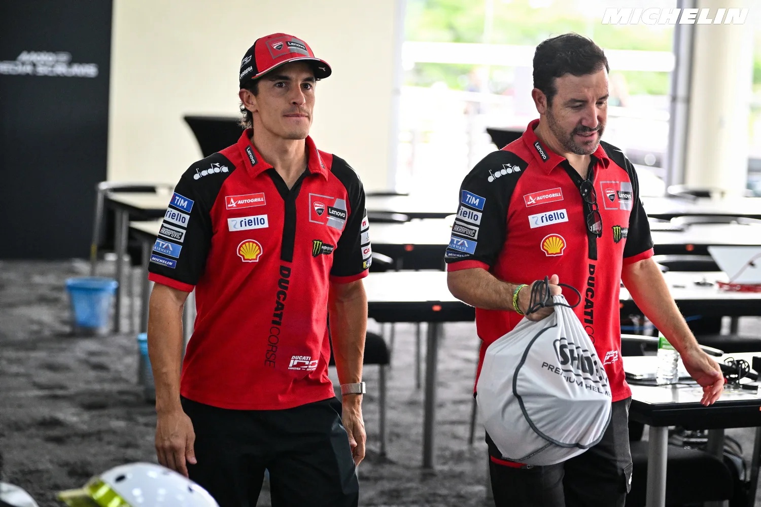 Rider in red Ducati Corse shirt and cap in the MotoGP paddock, walking past tables while a man carries a white bag; Michelin logo visible.