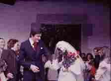 A bride in a white wedding dress and veil holds red flowers while walking with the groom in a dark suit and red tie, surrounded by smiling guests showering them with confetti