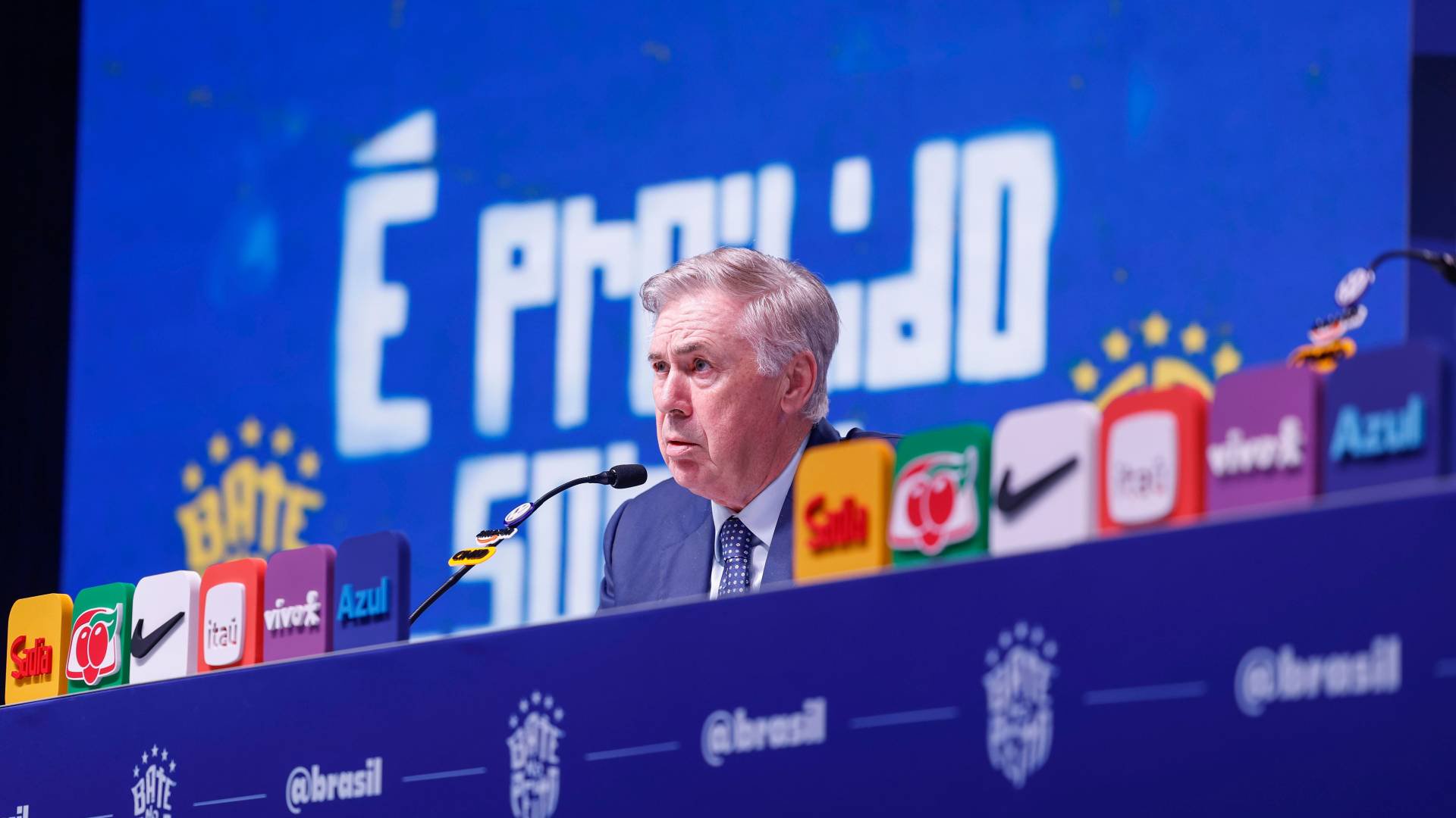 Homem de cabelos grisalhos, terno azul e gravata estampada, sentado em uma mesa azul com microfone, falando em coletiva de imprensa. Ao fundo, um telão azul com a frase É ORGULHO em branco e, na mesa, logotipos de patrocinadores em cores variadas