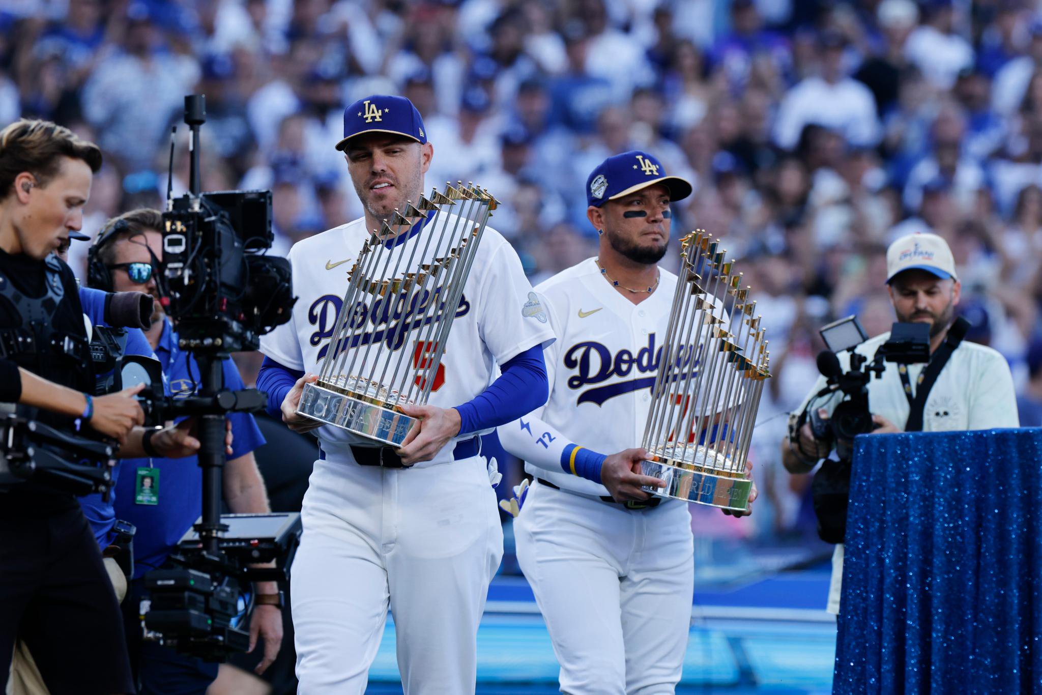 Two Los Angeles Dodgers players hold World Series trophies on the field, surrounded by cameras and fans.