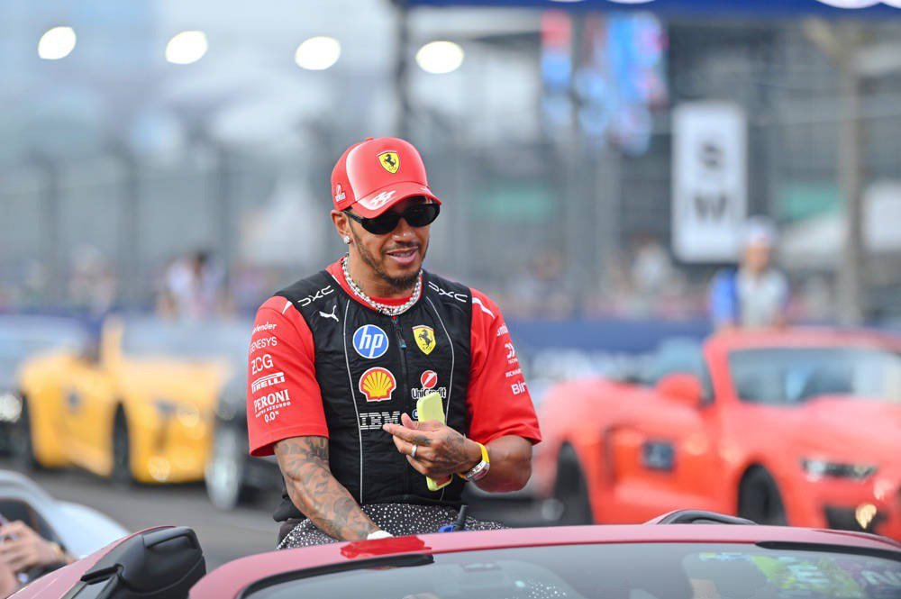 Driver in red Ferrari cap and sunglasses, wearing a Ferrari-branded race shirt with sponsor logos, riding in an open car at a Formula 1 event with spectators…