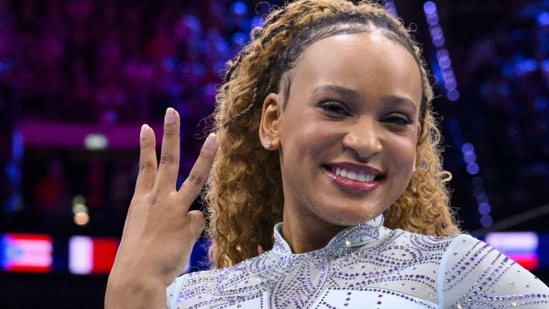 Ginasta negra sorrindo, cabelo cacheado preso, vestindo um collant branco com brilhos, levantando três dedos da mão esquerda, em um fundo desfocado de luzes roxas e azuis