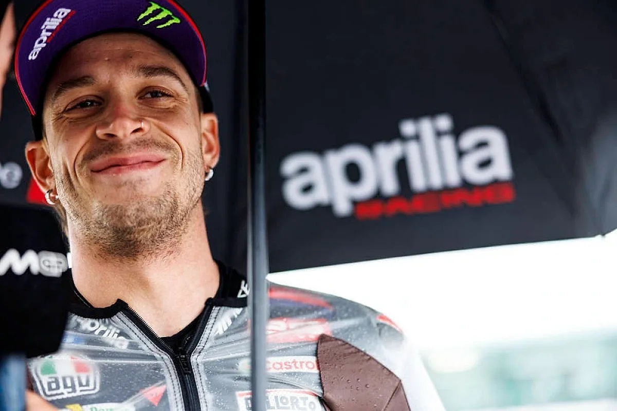 A rider in a grey Aprilia race suit and purple Monster-branded cap smiles beside an Aprilia Racing umbrella in the MotoGP paddock