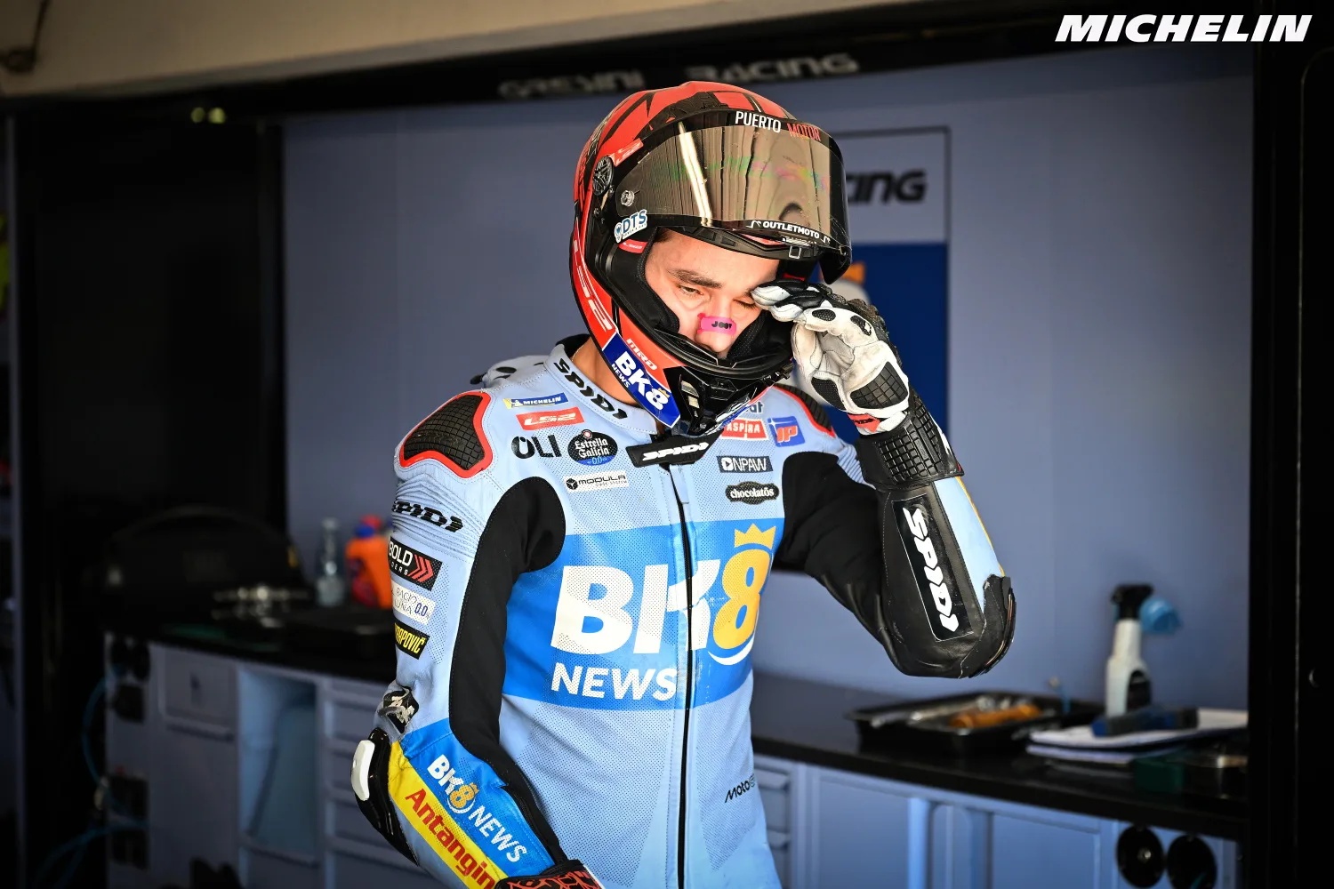 Rider in light blue and black leathers with BK8 News sponsor logos, wiping his face in the pit garage, Michelin branding visible