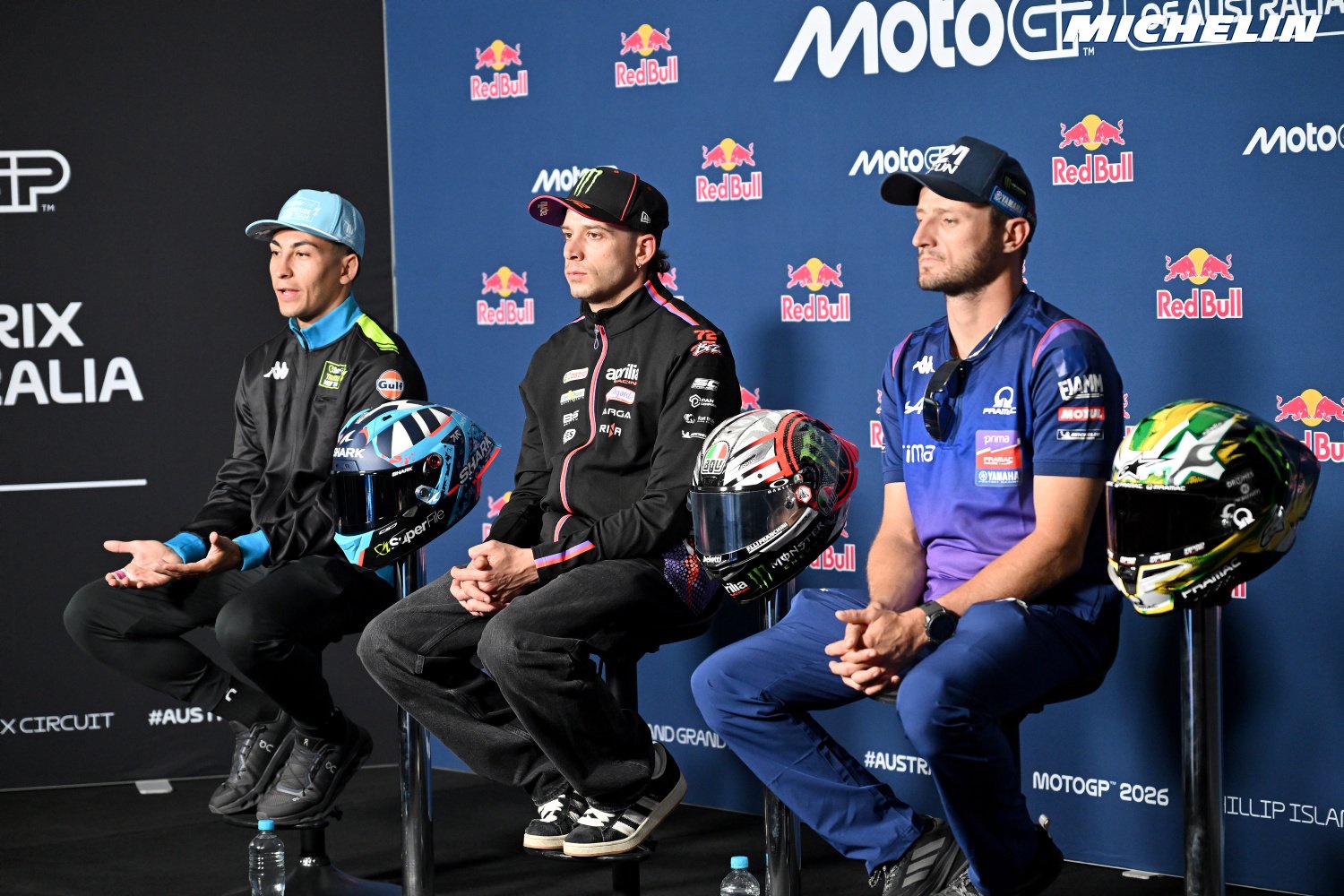 Three riders seated at a MotoGP press conference in Australia, with helmets beside them; the one on the left wears a light blue cap and black Gresini-style t…