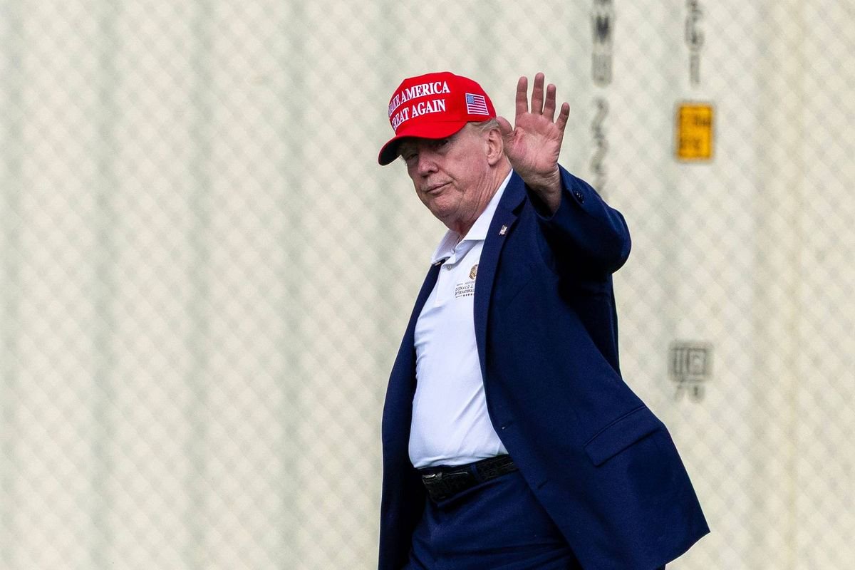 An individual dressed in a formal suit, including a white shirt and a navy blue blazer, is seen in mid-stride, extending a hand in a gesture of greeting or acknowledgment. The individual is wearing a red cap and is positioned against a backdrop of a light-colored, partially visible fence.