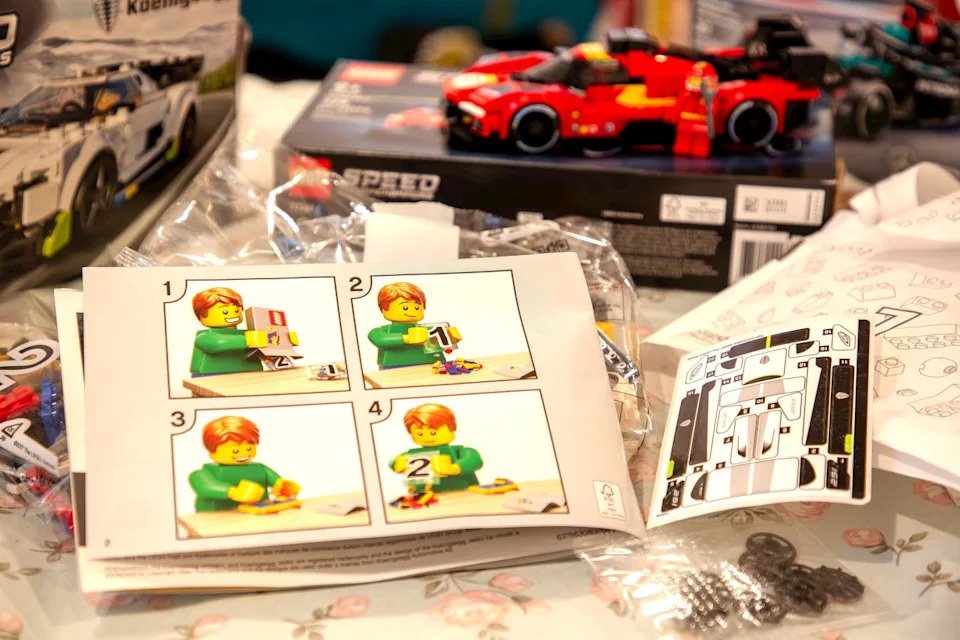Lego building instructions and pieces on a table with assembled car models in the background.