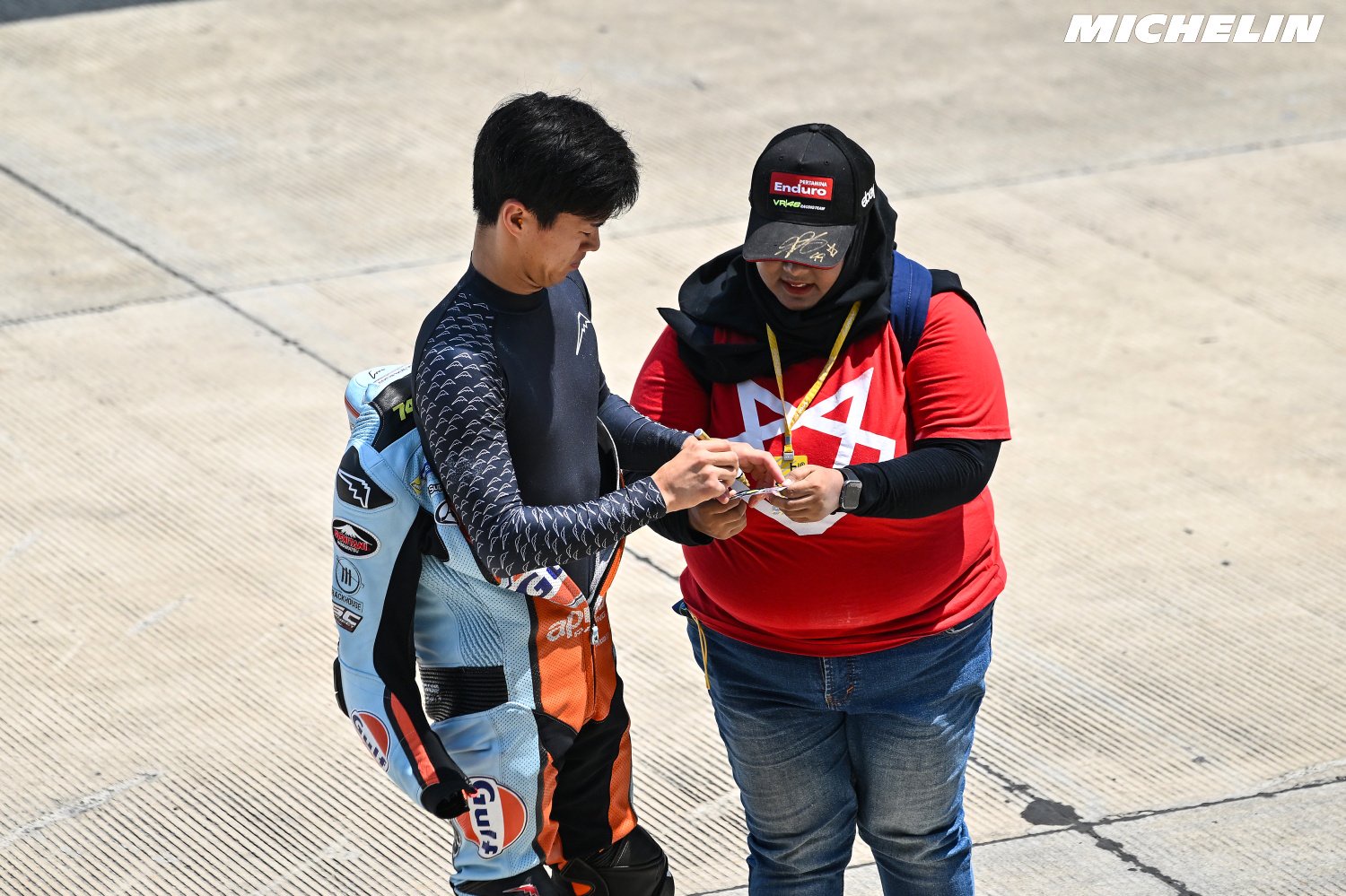 Rider in blue and orange race leathers with Ogura-related sponsor logos talks with a fan in a red shirt on a pit lane (Phillip Island)
